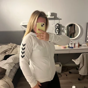 Vit hoodie från Hummel med dragkedja - Säljer en vit hoodie från Hummel med svarta chevron-detaljer på ärmarna och hel dragkedja framtill. Tröjan har huva, långärmad passform och är gjord i mjuk bomull. Perfekt för chill dagar eller träning. Snygg och sportig stil!