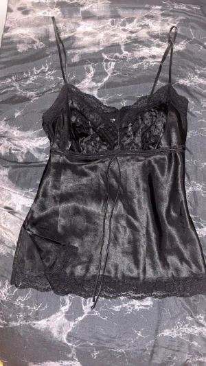 Svart satin linnetopp med spets - Supersnygg svart linnetopp i satin med tunna axelband och spetsdetaljer vid halsringning och nederkant. Toppen har en v-ringad form och dekorativ snörning framtill. Perfekt för dig som gillar en chic och lite edgy stil. Ena bandets grej på axeln har lossnat så går inte att justera den som vanligt.
