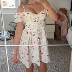 Vit blommig kortklänning med offshoulder - Supersöt vit kortklänning med Klänningen har offshoulder-ärmar och smala axelband, samt en figurnära midja (liten i storleken) Perfekt för sommarens alla tillfällen! 