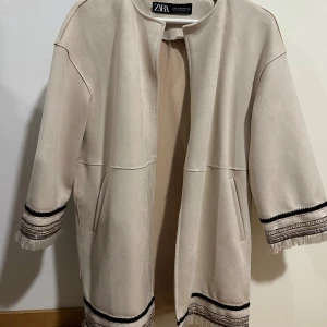 Beige kappa med fransar från Zara - Stilren beige kappa från Zara med snygga fransdetaljer och svarta dekorativa band vid ärmslut och nederkant. Jackan har rak passform, trekvartsärm och två fickor framtill. Perfekt för dig som gillar en clean look med unika detaljer.