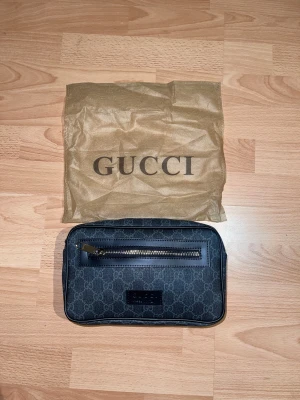 Svart axelväska från Gucci med GG-mönster - Snygg svart axelväska från Gucci med klassiskt GG-mönster över hela väskan. Väskan har en framficka med dragkedja och en bred axelrem i blått och rött. Materialet är skinn och polyester, och väskan har en stilren, rektangulär form.