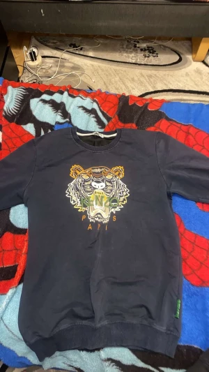 Mörkblå Kenzo sweatshirt med tiger - Säljer en mörkblå sweatshirt från Kenzo med ikonisk broderad tiger på bröstet i färgerna gul, vit och grön. Tröjan har rund hals, ribbade muddar och lång ärm. Perfekt för dig som vill sticka ut med ett coolt statement-plagg.