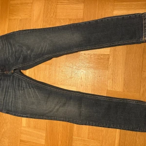 Blå jeans från Nudie Jeans, storlek 31/30 - Snygga blå jeans från Nudie Jeans i klassisk straight fit. Jeansen har fem fickor, orangea sömmar och subtila slitningar framtill. Materialet är mjukt denim och färgen är mörkblå med lätt tvättad look. Perfekt för dig som gillar stilrena och tidlösa jeans.