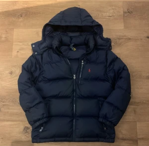 Mörkblå pufferjacka från Polo Ralph Lauren - Snygg mörkblå pufferjacka från Polo Ralph Lauren med huva och klassisk röd logga på bröstet. Jackan har dragkedja framtill, två sidofickor med dragkedja och quiltad design. Perfekt för kalla dagar och riktigt chill stil.