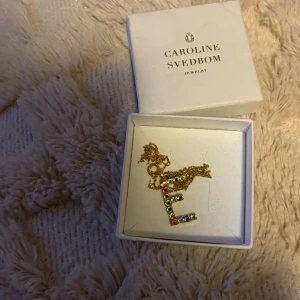 Caroline Svedbom halsband med E-berlock - Unikt halsband från Caroline Svedbom med en bokstavsberlock i form av E, dekorerad med gnistrande färgglada stenar. 