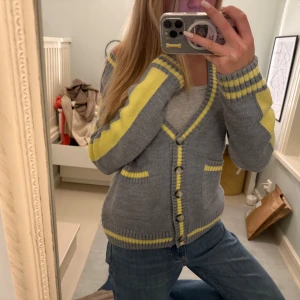 Grå kofta med gula detaljer - Säljer en grå stickad kofta med gula ränder och detaljer vid kanter, fickor och ärmar. Koftan har v-ringad hals, knappar framtill och två små fickor. Perfekt att slänga över en t-shirt för en chill look. 