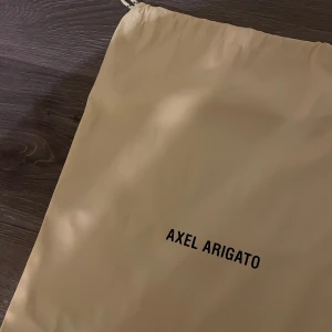 Beige Axel Arigato ryggsäck med snörning - Snygg beige ryggsäck från Axel Arigato med svart logga framtill. Väskan har praktisk snörning upptill och är tillverkad i ett mjukt syntetmaterial. Perfekt för dig som gillar minimalistisk stil och vill ha något enkelt att bära med dig tex träningskläder 