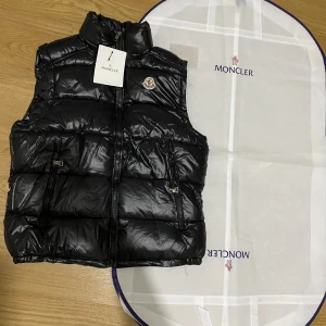 Svart dunväst från Moncler - Svart dunväst från Moncler med glansig finish och klassisk logotyp på bröstet. Västen har hög krage, två dragkedjefickor och quiltad design. Perfekt för lager-på-lager och snygg till streetwear. Dragkedja framtill och stilren look.
