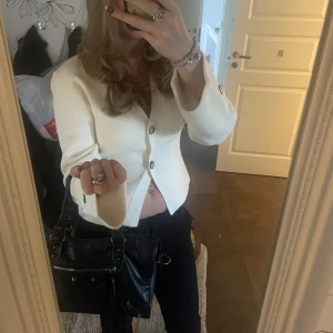 Vit stickad med knappar - Snygg vit stickad tröja med v-ringad hals och tre dekorativa knappar framtill. Tröjan har en stilren och modern look, perfekt att styla med jeans eller kjol. Materialet ser ut att vara mjukt och bekvämt, med en lätt struktur.