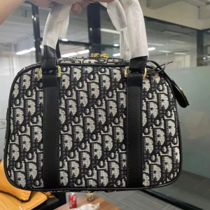 Svartvit handväska från Dior - Snygg handväska från Dior med ikoniskt monogrammönster i svart och vitt. Väskan har en boxig form, svarta skinn-detaljer och guldfärgade metalldelar. Materialet är en mix av textil och skinn, med två handtag och dragkedja upptill. Perfekt för dig som gillar lyxiga accessoarer med statement.