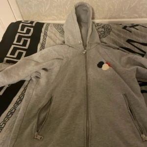 Grå Moncler hoodie med dragkedja - Säljer en grå hoodie från Moncler i storlek S. Tröjan har hel dragkedja, två fickor med dragkedja och en huva. På bröstet finns Monclers klassiska logomärke i svart, rött och vitt. Mjuk insida i bomullsmix, perfekt för chill dagar.