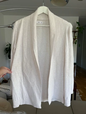 Beige ribbad kofta från MNG - Snygg beige kofta från MNG i storlek S. Koftan har ribbade detaljer längs kragen och muddar, öppen framtill och långärmad. Materialet är mjukt och stickat, perfekt för lager-på-lager under höst och vår. Enkel och clean look som funkar till allt.