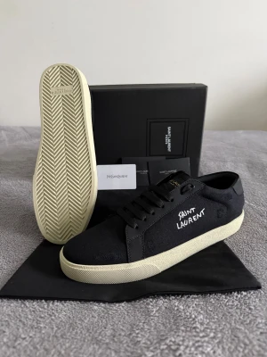Saint Laurent skor - Svarta sneakers från Saint Laurent med vit broderad logga på sidan. Skorna har en klassisk låg profil, svarta snören och vit, texturerad gummisula. Ovandelen är i canvas och detaljer i skinn. Perfekt för dig som gillar stilrena och exklusiva sneakers.