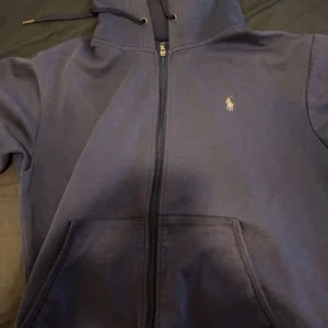 Mörkblå hoodie från Ralph Lauren - Säljer en snygg mörkblå hoodie från Ralph Lauren i bra skick. Den har en dragkedja framtill och en liten broderad logga på bröstet. Perfekt för höst och vår, med en skön passform och praktiska fickor. Perfekt för en casual look! Storlek S. OBS enda defekten är att metall grejen på ena snöret har lossnat.