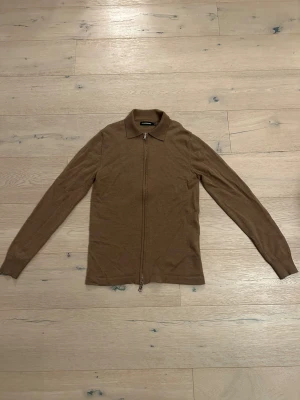 Brun kofta med dragkedja från J.Lindeberg - Snygg brun kofta från J.Lindeberg med hel dragkedja framtill och klassisk krage. Långärmad och tillverkad i mjukt material. Perfekt för lager-på-lager och stilrena outfits. Den är i storlek S men passar även XS.