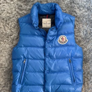 Blå dunväst från Moncler - Snygg blå dunväst från Moncler med hög krage och ikonisk logga på bröstet. Västen har två dragkedjefickor fram och knäppning med tryckknappar. Insidan är svart och glansig, och materialet är syntet med dunfyllning. Perfekt för att addera lite lyx till din outfit.