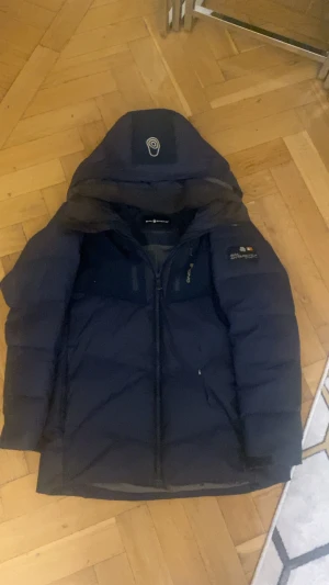 Mörkblå Sail Racing dunjacka - Säljer en mörkblå dunjacka från Sail Racing med huva och snygga detaljer. Jackan har broderad logga på ärmen, dragkedja framtill och två sidofickor. Insidan är grå och jackan är quiltad för extra värme. Perfekt för kalla dagar.