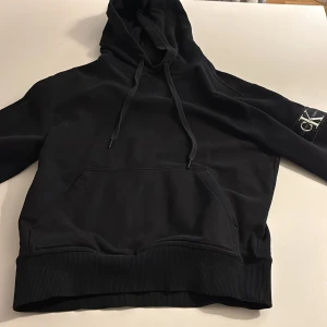 Svart hoodie Calvin Klein Jeans XS - Svart hoodie från Calvin Klein Jeans i storlek XS. Tröjan har en stor CK-logga på ärmen, huva med snörning och en klassisk magficka. Ribbad nederkant och ärmslut ger en snygg passform. Perfekt för dig som gillar stilrena streetwear-plagg.