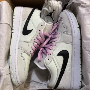 Nike Air Jordan 1 Low SE med rosa snören - Nike Air Jordan 1 Low SE sneakers i ljusgrön och vit färg med svarta swoosh-loggor på sidorna. Skorna har platt sula, rund tå och perforerat tåparti. De är tillverkade i skinn och textil, och har snygga rosa snören samt extra vita snören med Jumpman-logga.