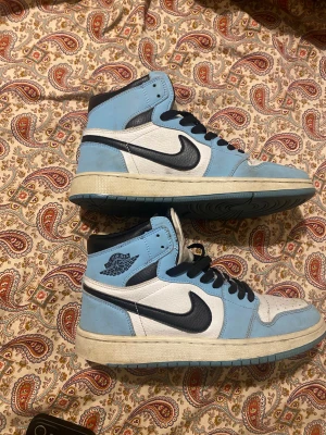 Nike Air Jordan 1 High blå/vit - Säljer ett par Nike Air Jordan 1 High sneakers i blått och vitt med svarta detaljer. Skorna har klassisk snörning, rund tå och platt sula. Ovandelen är i skinn och mocka, med perforerad tåbox och ikonisk Jordan-logga på sidan. Perfekt för dig som gillar streetwear och sneakers.