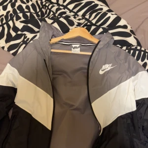 Nike vindjacka i grått och vitt - Nike vindjacka med huva i snygg grå, vit och svart färgblock-design. Jackan har dragkedja framtill och klassisk Nike-logga på bröstet. Perfekt för att hålla vinden borta och samtidigt se sportig ut.