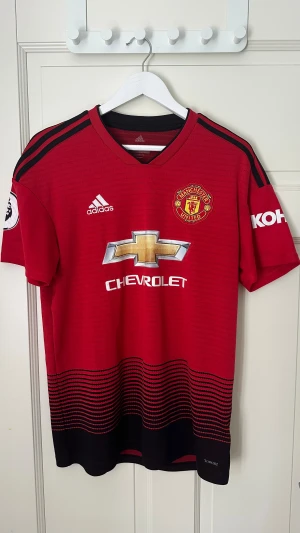 Äkta manchester united fotbollströja 18/19 M - Manchester Uniteds officiella matchtröja från Adidas med Premier League-märke på ärmen. Röd med svarta detaljer, svarta ränder på axlarna och vågigt mönster nertill. Broderat klubbmärke och Chevrolet-logga på bröstet. Tillverkad i lätt och ventilerande polyester. Väldigt bra skick 