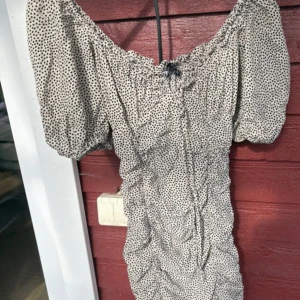 Prickig kortklänning med puffärm XS - Supersöt kortklänning med puffiga kortärmar och offshoulder-modell. Klänningen är beige med svarta prickar och har rynkade detaljer längs hela plagget. Perfekt för dig som gillar en feminin och trendig look.