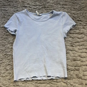 Ljusblå ribbad topp från Nelly - Säljer en ljusblå ribbad topp från Nelly med smala vertikala ränder. Toppen har rund halsringning, korta ärmar och vågig kant vid ärmslut och nederkant. Perfekt för dig som gillar enkel och fräsch stil.
