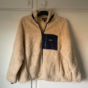 Beige Pile Jacka från Patagonia | M - Mysig beige teddyjacka från Patagonia med marinblå detaljer och bröstficka med dragkedja. Jackan har hög krage, hel dragkedja framtill och är tillverkad i fluffig fleece. Perfekt för kalla dagar och riktigt skön att bära.
