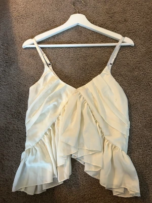 Vit omlott linnetopp med volanger - Säljer en ljusvit linnetopp med omlott-design och volanger. Toppen har smala justerbara axelband och är tillverkad i ett lätt, flödande material som ger en snygg och luftig känsla. Perfekt för dig som gillar romantiska detaljer och en feminin stil.