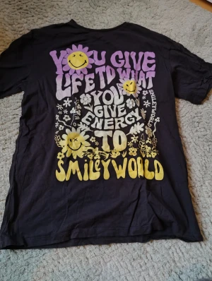 Svart SmileyWorld t-shirt från H&M - Cool svart t-shirt från H&M x SmileyWorld med färgglad print i lila, vitt och gult på magen. Trycket har glada smileys, blommor och texten 'You give life to what you give energy to'. Skön oversized passform och mjukt bomullsmaterial, perfekt för dig som gillar statement-plagg.