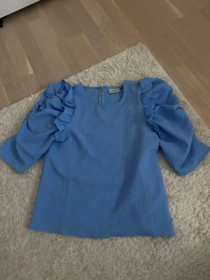 Blå blus med volangärmar från ZINK - Säljer en blå blus från ZINK i storlek XS. Blusen har puffiga trekvartsärmar med volangdetaljer och är tillverkad i ett strukturerat syntetmaterial. Rund halsringning och rak passform ger en clean look. Perfekt för dig som gillar statement-ärmar.