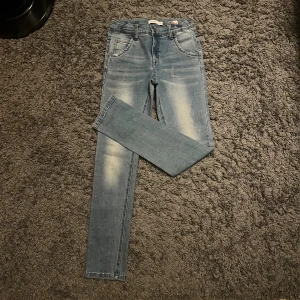 Blå jeansbyxor från Name It, strl 146 - Snygga ljusblå jeansbyxor från Name It med klassisk femficksdesign och coola slitningar framtill. Modellen är rak med normal passform och har detaljerade bakfickor med broderad logga. Perfekt för dig som gillar en avslappnad streetstil. Perfekt för 11-12 år. Storlek 146
