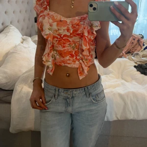 Blommig croppad linnetopp från H&M - Superfin croppad linnetopp från H&M i storlek XS. Toppen har ett blommigt mönster i orange, rosa och vitt, volangdetaljer framtill och på axlarna samt en omlottliknande känsla. Perfekt för sommarens alla häng och ger en fräsch vibe.