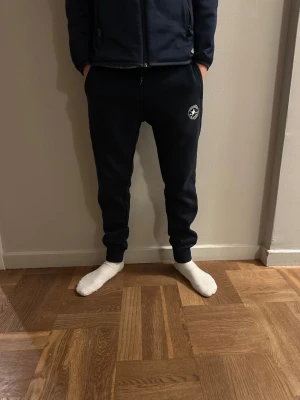 Mörkblå mjukisbyxor från Converse märket är lite söner  - Snygga mörkblå mjukisbyxor från Converse med klassisk All Star-logga på benet. Byxorna har ribbade muddar vid benslut, snörning i midjan och är gjorda i mjukt bomullsmaterial. Perfekt för chill dagar eller träning.