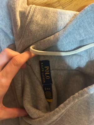 Grå hoodie från Polo Ralph Lauren - Säljer en grå hoodie från Polo Ralph Lauren i storlek S. Tröjan har huva med snörning, lång ärm och en klassisk känguruficka framtill. Materialet är mjuk bomull och polyester, perfekt för chill dagar. Diskret Polo-logga broderad på bröstet.