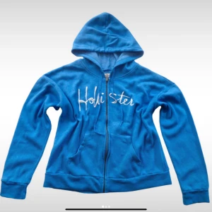 Blå hoodie från Hollister med dragkedja - Snygg blå hoodie från Hollister med vit broderad logga på bröstet. Tröjan har huva, långa ärmar och praktisk dragkedja framtill. Mjuk och skön, perfekt för chill dagar eller när du vill ha en avslappnad stil.