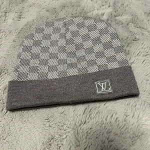 Grå rutig mössa från Louis Vuitton - Snygg grå mössa från Louis Vuitton med klassiskt rutigt mönster och LV-logga framtill. Mössan är stickad och har en uppvikt kant för extra stilpoäng. Perfekt accessoar för att lyfta din vinterlook.