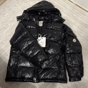 Svart Moncler Maya pufferjacka med huva - Säljer en svart pufferjacka från Moncler med glansig finish, huva och dragkedja framtill. Jackan har klassisk Moncler-logga på ärmen och coolt serietidningsfoder på insidan. Perfekt för dig som vill ha en varm och snygg vinterjacka med streetkänsla.