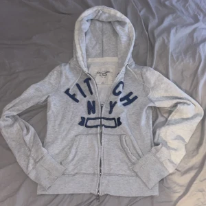 Grå hoodie Abercrombie & Fitch xs/s - Snygg ljusgrå hoodie från Abercrombie & Fitch i storlek M men passar mig som har xs/s Tröjan har blå broderad text på bröstet, två fickor fram och en rejäl huva. Dragkedjan har A&F-logga och materialet är mjukt bomullsmix. Perfekt för chill dagar och streetstyle.