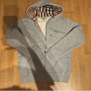 Grå Burberry hoodie med dragkedja - Säljer en grå hoodie från Burberry med klassiskt rutigt foder i huvan och insidan. Tröjan har dragkedja framtill, snörning vid huvan och två fickor. Perfekt för dig som vill ha en stilren och lyxig look med ikoniskt Burberry-mönster. Sitter däremot mer m storlek