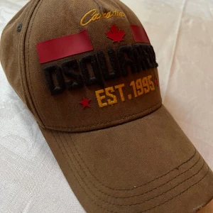 Dsquared2 keps brun - Säljer en brun keps från Dsquared2 med broderad text, röda detaljer och gul brodyr. Framsidan har tydlig Dsquared2-logga, kanadensiskt lönnlöv och EST. 1995 i gult. Kepsen har böjd skärm och klassisk passform. Perfekt för dig som vill sticka ut med en snygg accessoar.