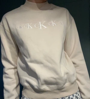 Beige sweatshirt från Calvin Klein Jeans - Snygg beige sweatshirt från Calvin Klein Jeans med broderad CK-logga framtill. Tröjan har ribbad hög krage, muddar vid ärmslut och nederkant samt långärmad passform. Perfekt för dig som gillar stilrena och klassiska plagg med en modern touch.