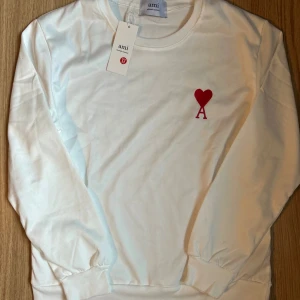 Vit sweatshirt från Ami Paris med hjärtdetalj - Vit sweatshirt från Ami Paris med röd broderad hjärtdetalj och bokstaven A på bröstet. Klassisk rund halsringning, långa ärmar och ribbade muddar. Tillverkad i mjuk bomull för en skön och avslappnad känsla.
