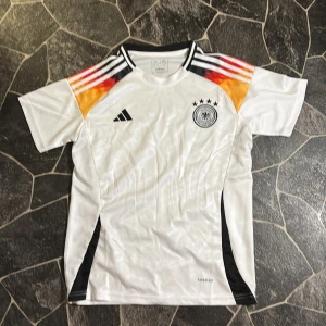 Tyskland fotbollströja Adidas - Säljer en officiell Tysklands fotbollströja från Adidas i storlek L. Tröjan är vit med svarta, röda och gula detaljer på axlarna, samt svarta ränder och emblem på bröstet. Materialet är lätt och andas, Aeroready-teknologi. Klassisk rund hals och korta ärmar.