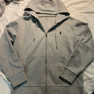 Grå hoodie från Polo Ralph Lauren - Säljer en grå hoodie från Polo Ralph Lauren med dragkedja, huva och snörning. Tröjan har långa ärmar, två fickor fram och den klassiska Polo-loggan broderad på bröstet. Perfekt för chill dagar och enkel att matcha med jeans eller joggers.