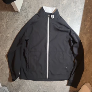 Svart vindjacka från FootJoy - Svart vindjacka från FootJoy med vit dragkedja och logga på kragen och ryggen. Jackan har hög krage, två framfickor och är långärmad. Perfekt för dig som gillar sportig stil och vill ha något lätt och funktionellt.
