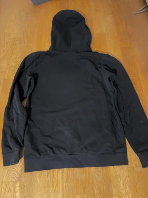 Svart hoodie från Peak Performance - Svart hoodie från Peak Performance med stor vit logotyp på bröstet. Tröjan har huva, ribbade muddar och en klassisk magficka framtill. Perfekt för chill eller träning, enkel och stilren design som funkar året runt.