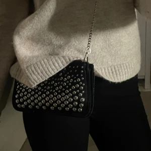 Svart kuvertväska med silvernitar - Svart kuvertväska med massor av silverfärgade nitar på framsidan. Väskan har en avtagbar kedja i silvermetall och stängs med dragkedja. Perfekt storlek för mobilen och det viktigaste. I mocka immitation.
