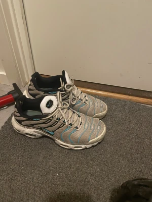 Nike Air Max Plus grå/blå sneakers - Nike Air Max Plus sneakers i grå mesh med blå och svarta detaljer. Snyggt vågigt mönster på ovansidan och vit sula med synliga luftkuddar. Skorna har snörning och rund tå, samt svart insida och vit etikett med blå swoosh på plösen.